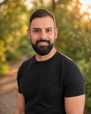 Portrait of a bearded man wearing a black t-shirt smiling at the camera with blurred green foliage in the background